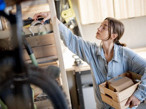 Young handywoman searching some working tools on a wooden shelves in the workshop. Concept of organization in home workshop or storage
